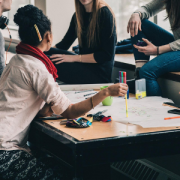 woman sitting at work table; team members standing/sitting on nearby desks