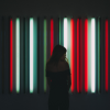 Person in hoodie in front of red white black and green vertical neon tubes that look like barcode